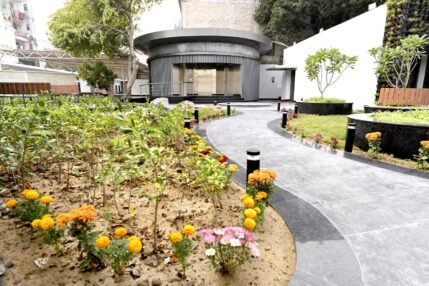 Landscaped garden pathway with flowering plants leading to the Sulabh Aspirational Toilet building.